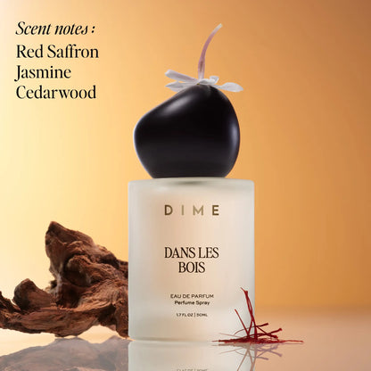 Black and white perfume bottle labeled "Dans Les Bois" with purple ribbon bow, surrounded by wood and saffron threads on tan 