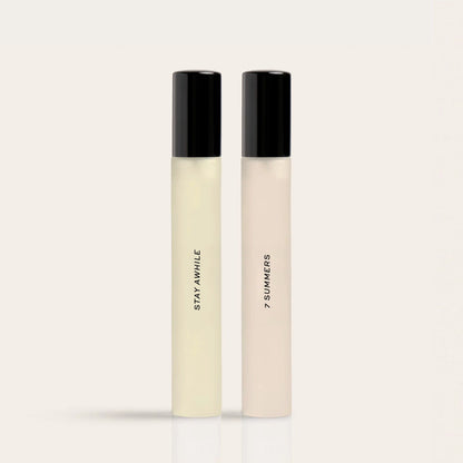 Two cylindrical fragrance bottles with black caps, one containing pale yellow liquid labeled "Stay Awhile" and one with beige