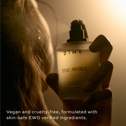 Cream-colored perfume bottle labeled "Stay Awhile" by Dime held in hand against warm, golden lighting.